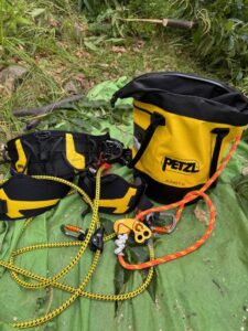 Climbing gear spread on a green tarp: yellow Petzl bag, black‑yellow harness, two ropes (yellow and orange), carabiners and a belay/descender device ready for use.