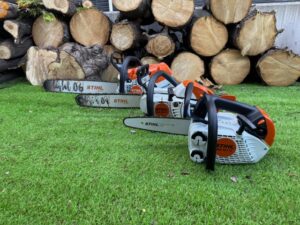 Three STIHL chainsaws with orange and gray bodies lie on a green lawn in front of a stacked log pile.