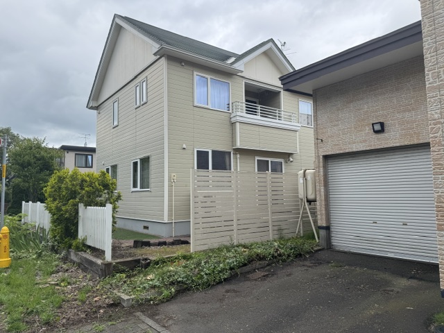 Two-story beige house with a small balcony, fenced front yard, and a neighboring brick garage with a roller door under a cloudy sky.