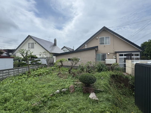 Front yard of a suburban home with trimmed shrubs, a rock garden, and a low stone wall, two gable-roof houses in the background under a cloudy sky.