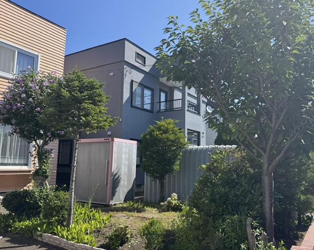 Two-story gray apartment building with black-framed windows, flanked by a pink-trim storage shed and lush garden under a blue sky.