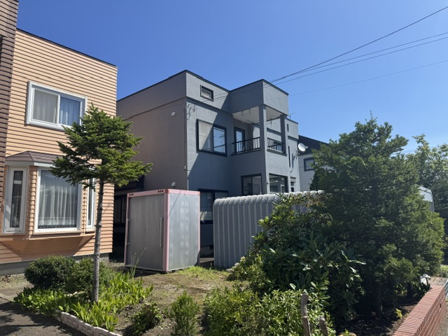Front view of a modern gray multi-unit building with balconies, next to a peach-colored wooden house, with small garden and storage shed in the yard under a blue sky.